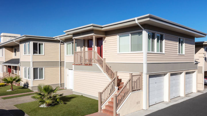 A modern two-story apartment building with beige siding, red doors, and a well-kept lawn featuring small palm trees in the front.