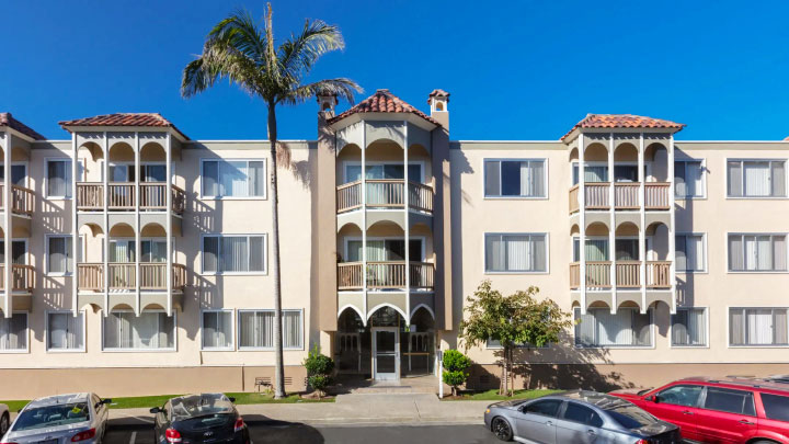 Front view of a beige apartment building with balconies, large windows, a central entrance, and palm trees. Cars parked in front.