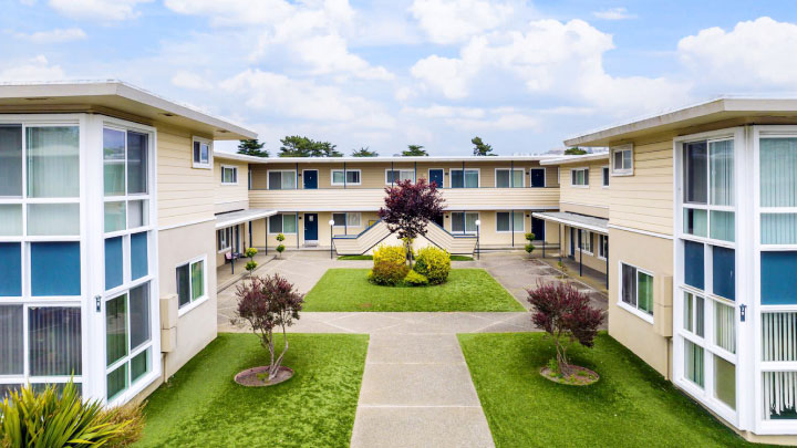 A two-story apartment complex with beige siding, blue doors, exterior walkways, and a central courtyard featuring green lawns and small trees.