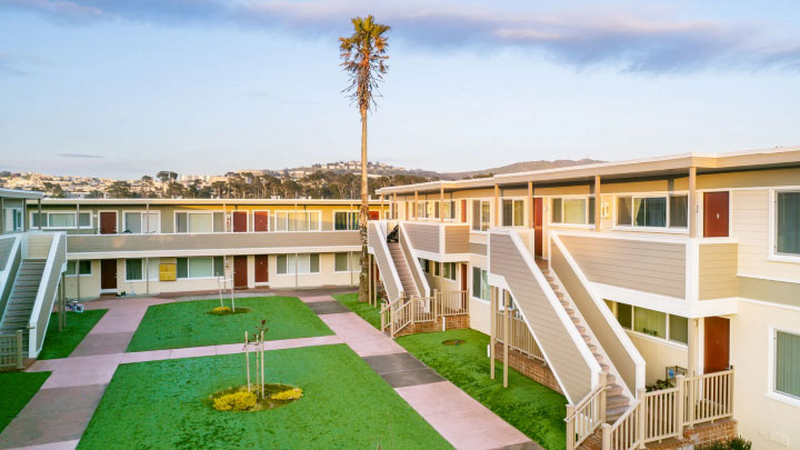 A modern two-story apartment complex with beige and white siding, exterior staircases to the second floor, and well-maintained green lawns with palm trees in the background.