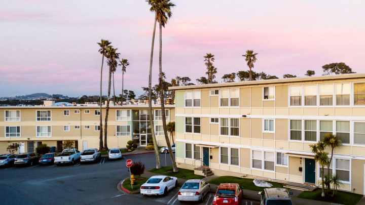 Beige and white apartment building with windows and palm trees. Cars are parked in front, with a stop sign in the lot.